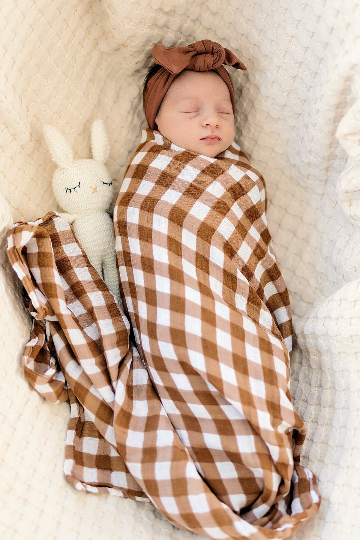 A baby wrapped in a brown and white gingham muslin swaddle blanket, lying with a knitted bunny toy.