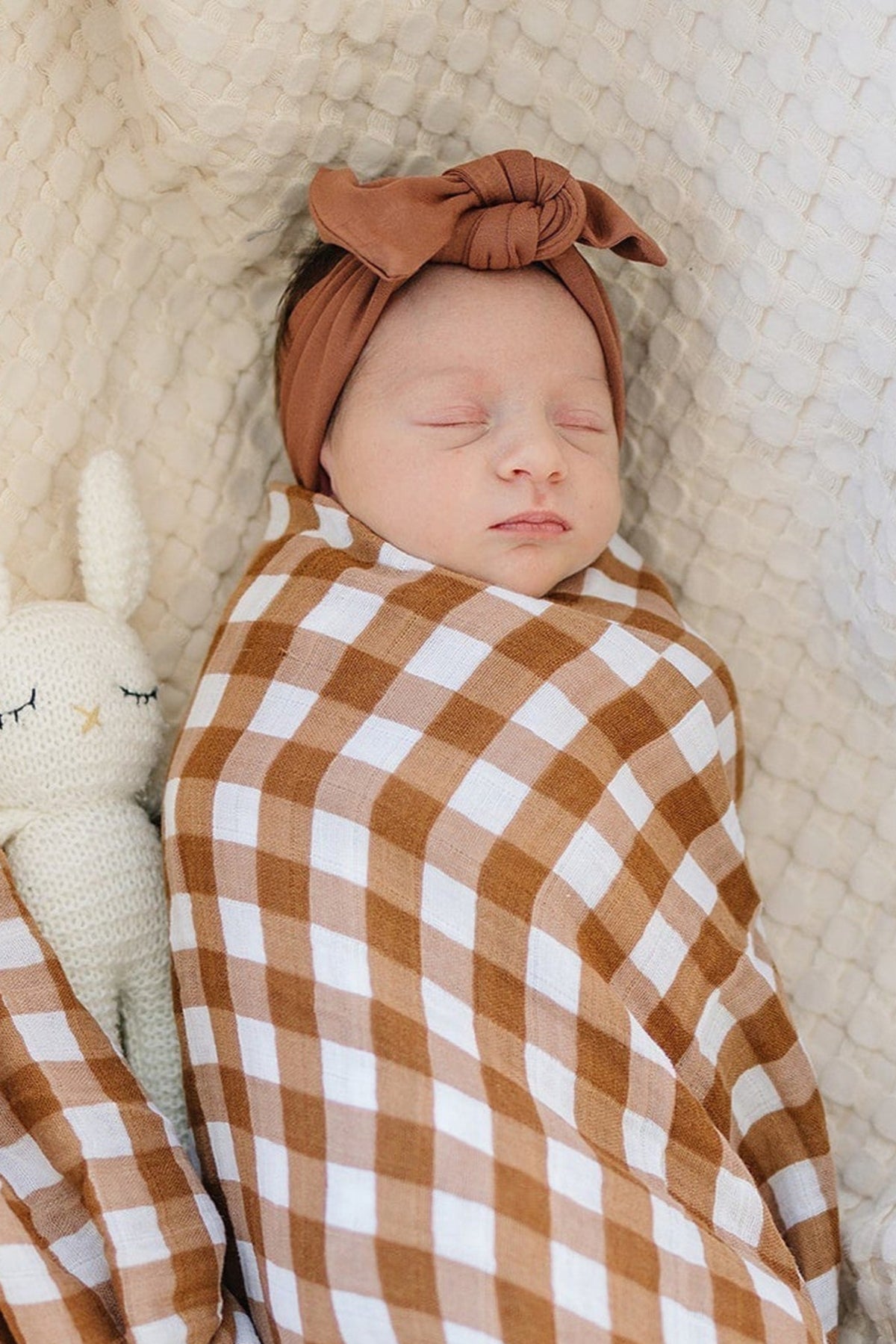 A sleeping baby in a rust head wrap and brown checkered blanket, next to a knitted white rabbit toy.