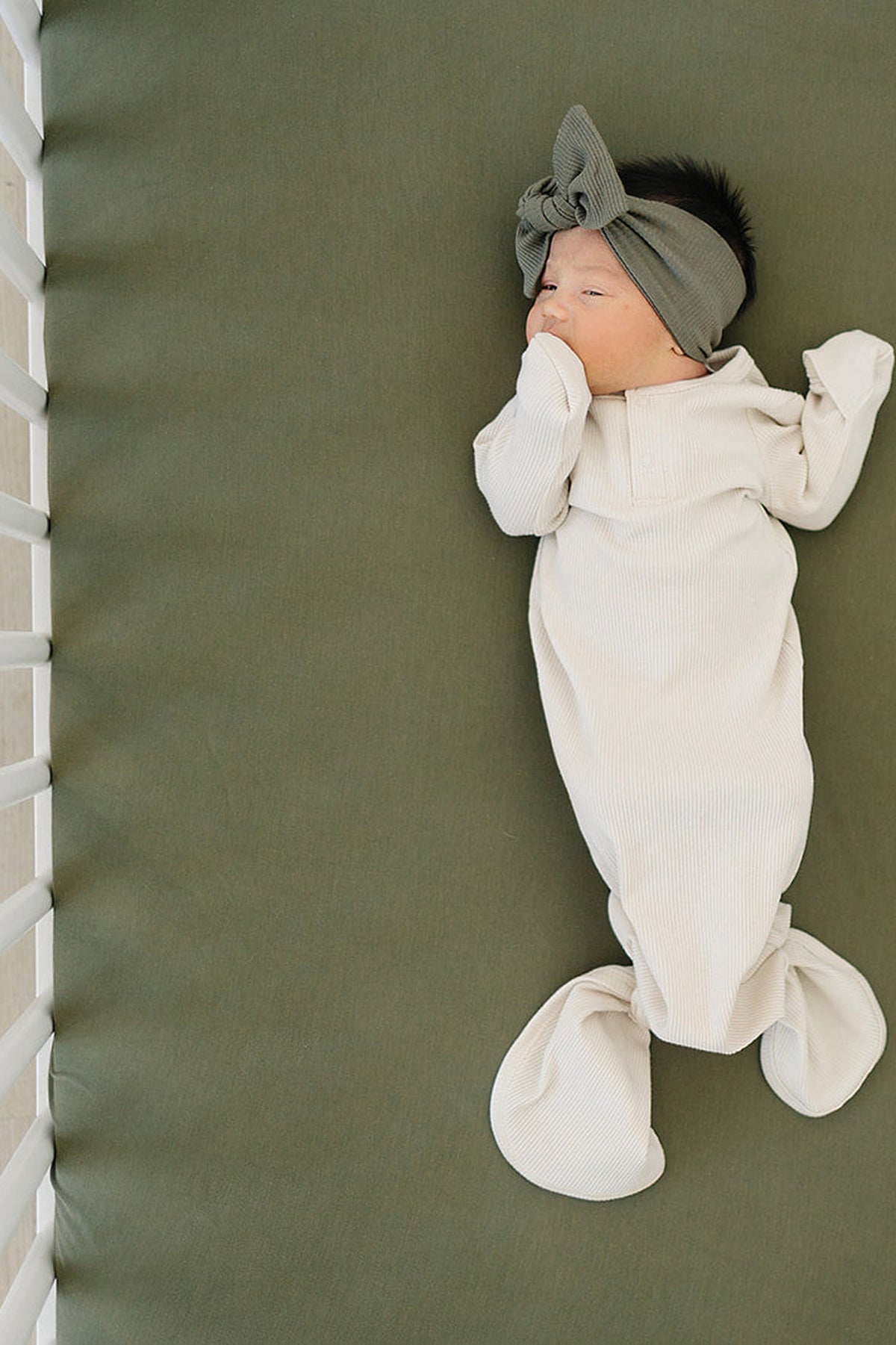 A baby in a cream outfit and green headband lies on an olive crib sheet in a white crib.