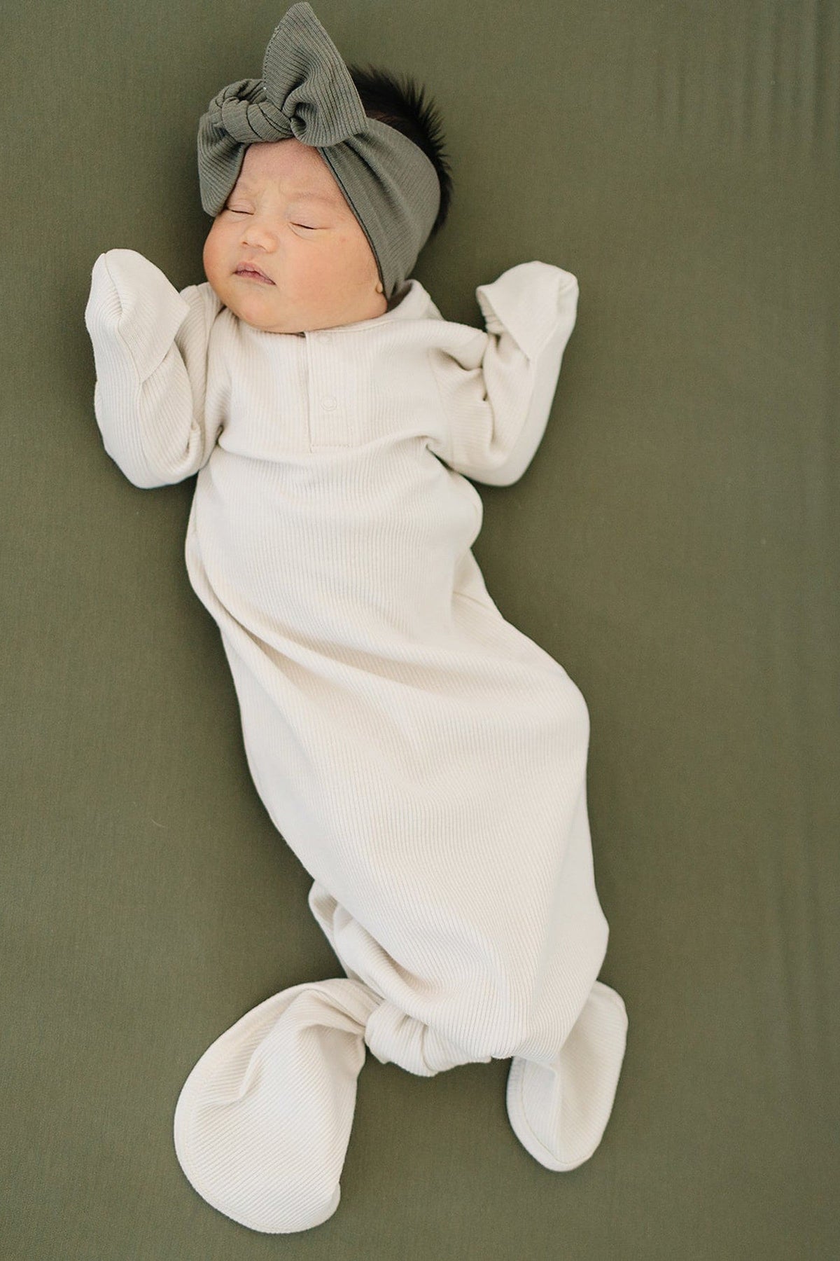 A baby in a vanilla ribbed knot gown and a grey headband lies on a green surface.