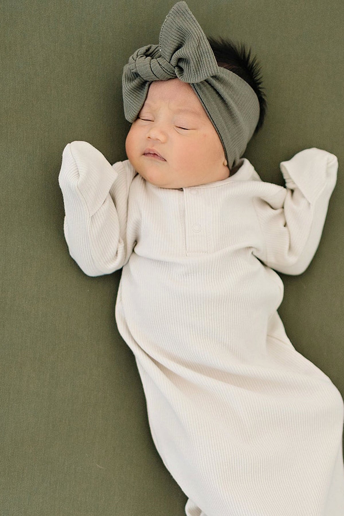 A baby wearing a green ribbed head wrap and white ribbed outfit lies on a green surface.