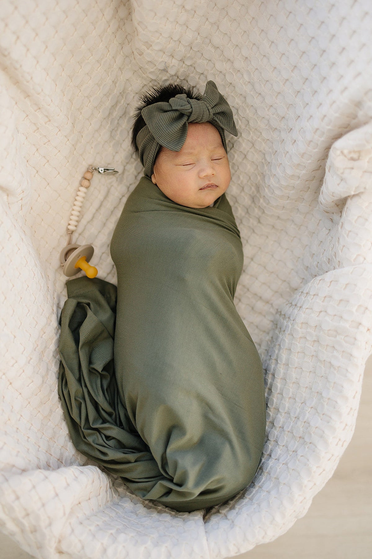 A baby wrapped in an olive stretch swaddle blanket lies in a cozy basket, wearing a matching headband.