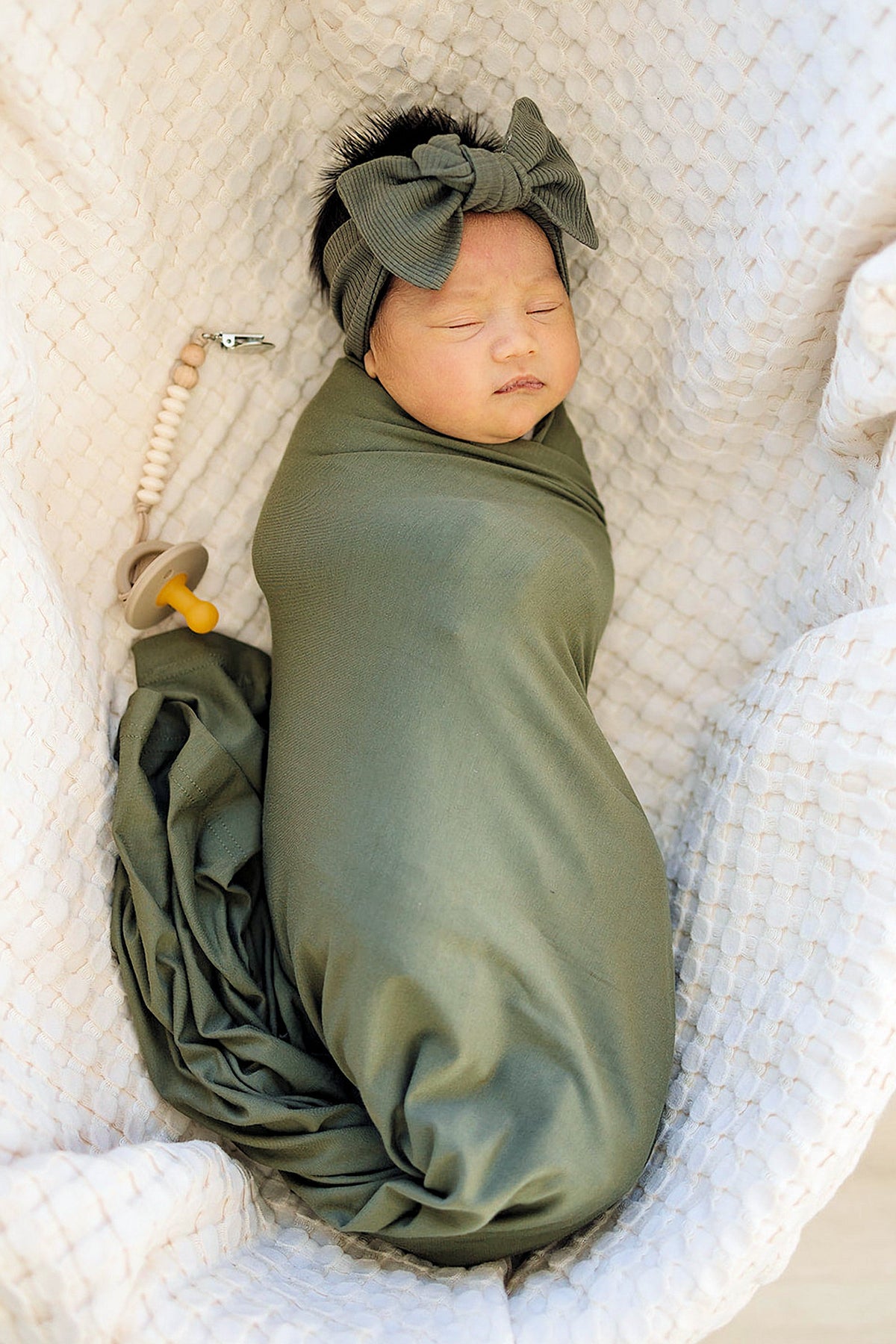 A baby swaddled in green fabric with a matching ribbed head wrap, resting on a textured white blanket.