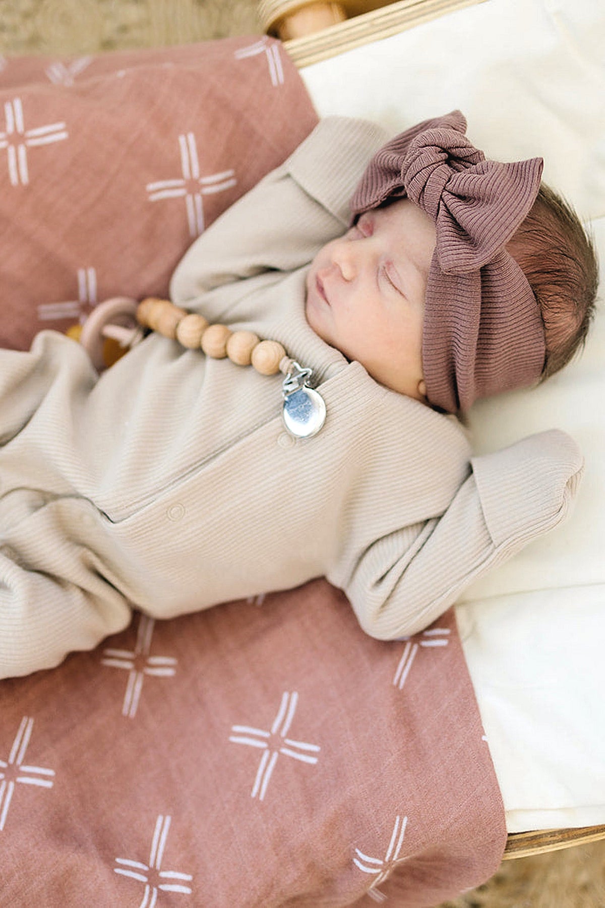 Newborn in oatmeal ribbed pajama and mauve headband, lying on patterned blanket with wooden beads.