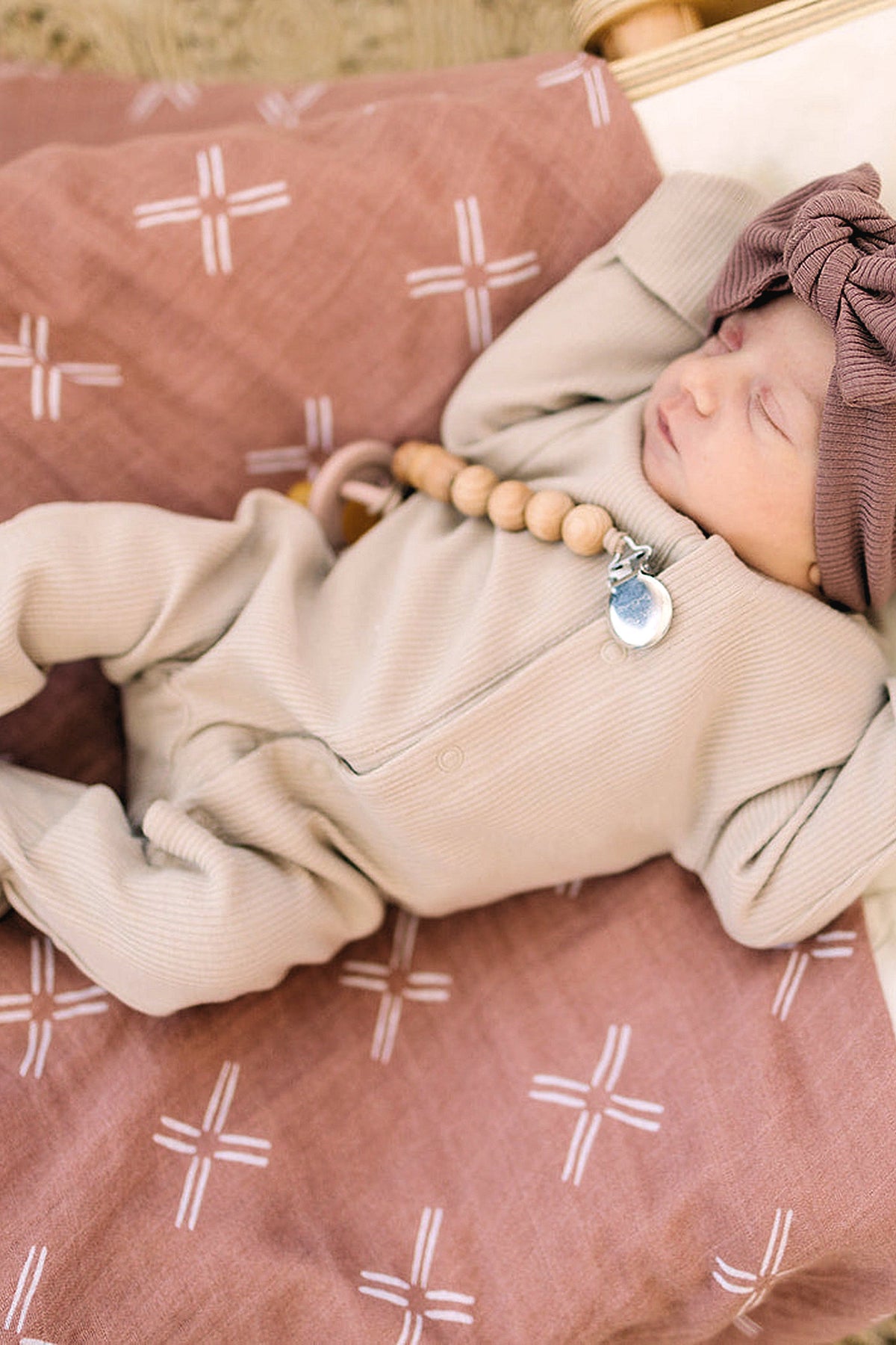 A baby sleeps on a patterned peach muslin blanket, wearing a beige outfit and matching headband, with a wooden bead.