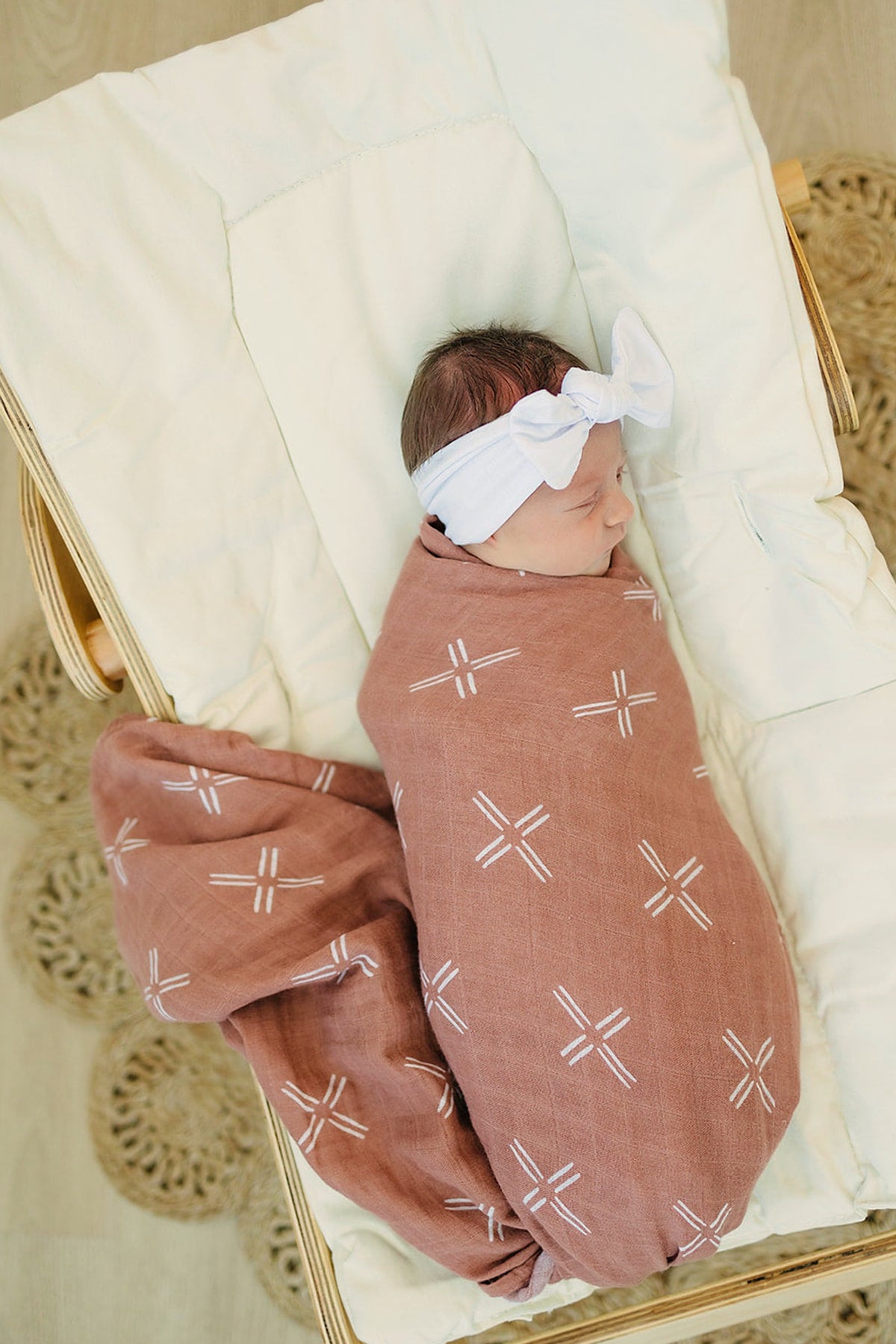 A baby wrapped in a peach-colored muslin swaddle blanket with white patterns, lying in a cushioned basket.