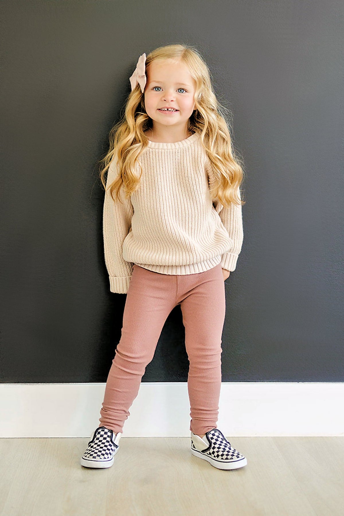 A child wearing dusty rose leggings and a cream sweater stands against a black wall, smiling.