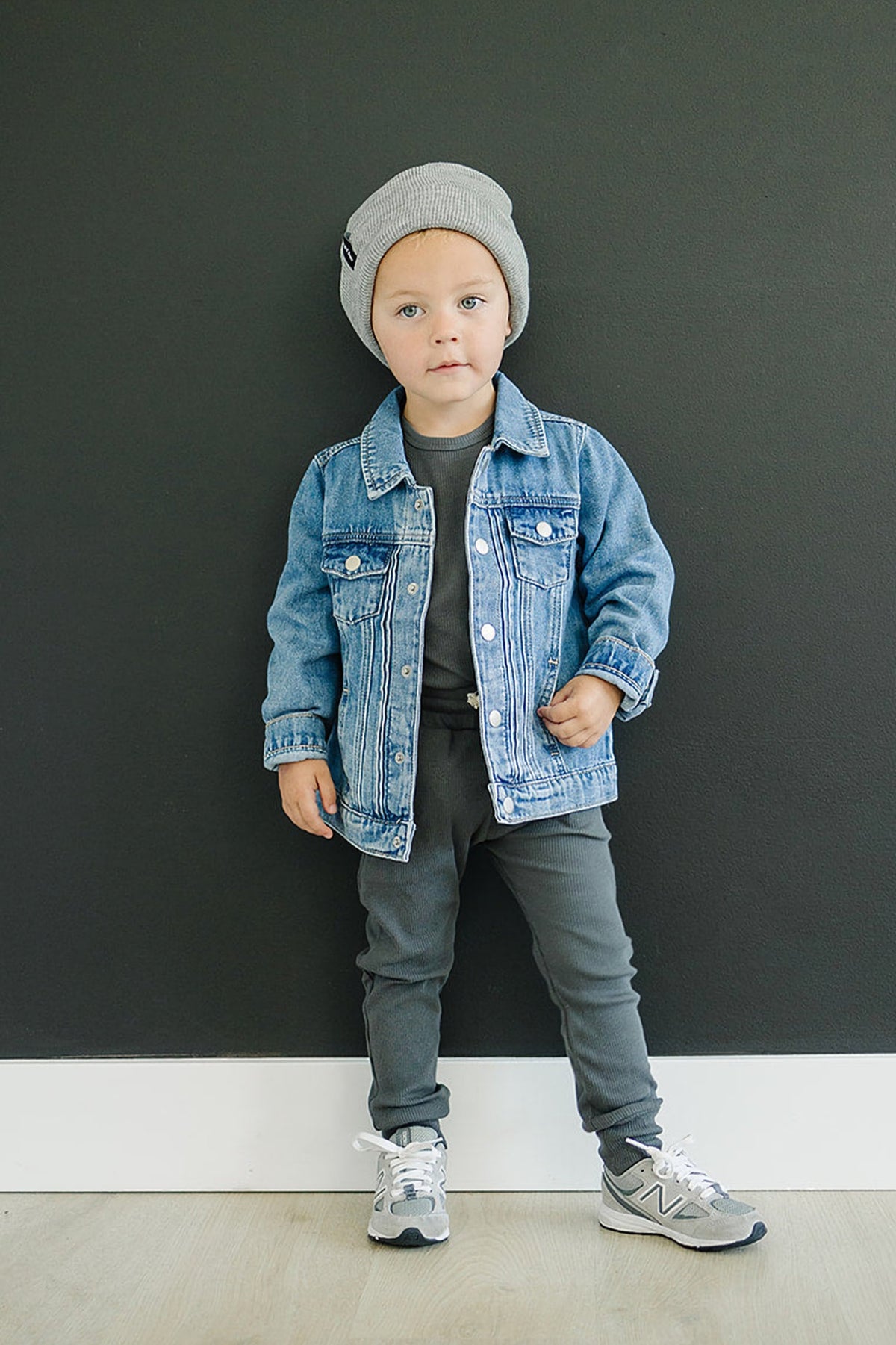 A toddler wearing a blue denim jacket, gray beanie, and gray pants stands against a dark background.
