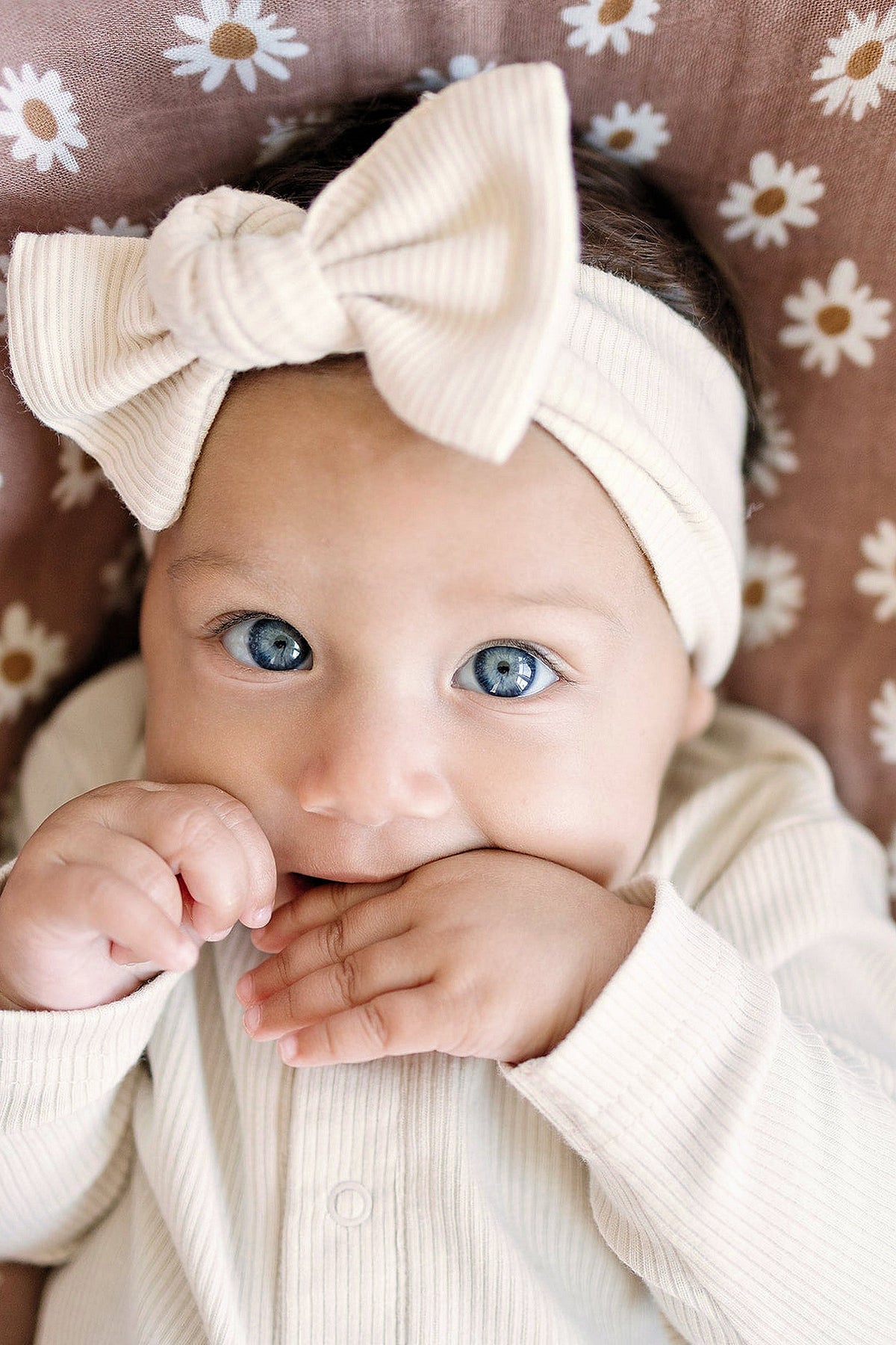 Baby wearing a vanilla ribbed head wrap with a bow, lying on a daisy-patterned blanket.