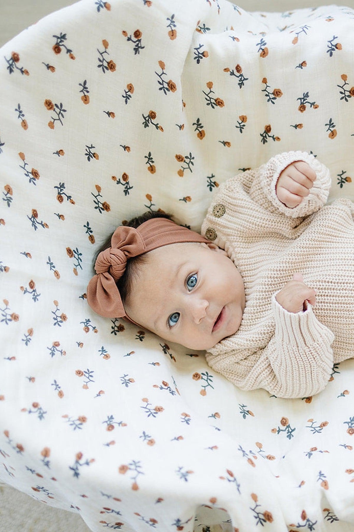Baby wearing a rust-colored head wrap and beige knitted sweater lies on a floral-patterned blanket.