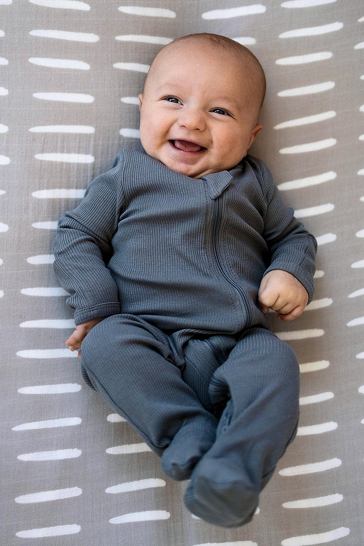 Smiling baby wearing charcoal ribbed footed pajamas, lying on a patterned gray blanket with white stripes.