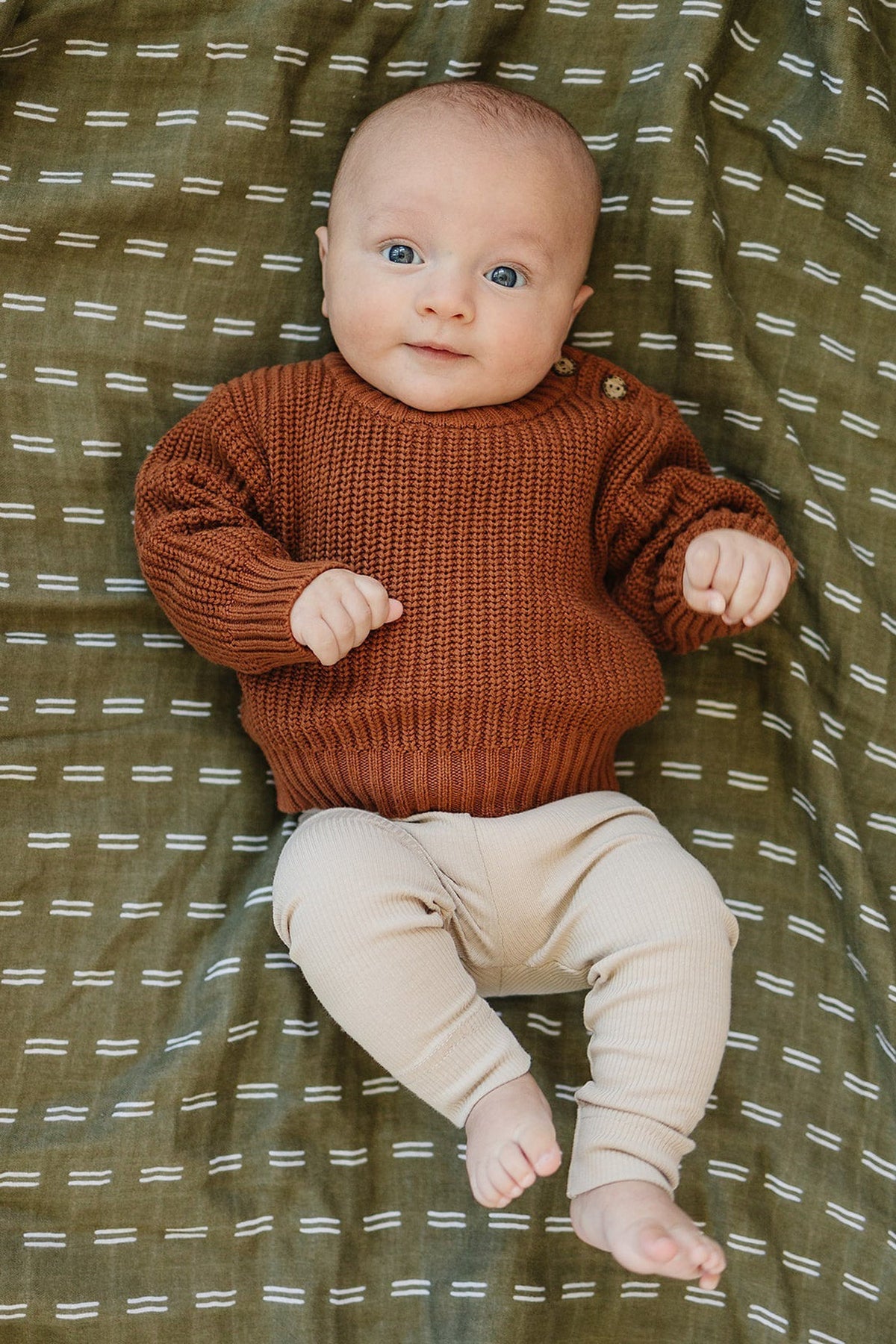 Baby wearing oatmeal-colored leggings and a rust sweater lying on a patterned green blanket.