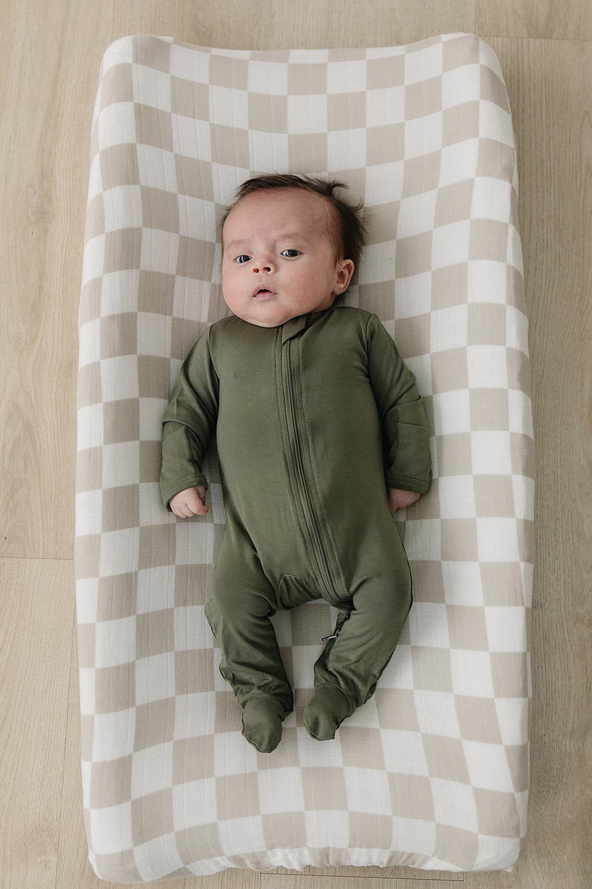 A baby lies on a taupe checkered muslin changing pad cover, wearing a green zip-up onesie. The soft, breathable fabric complements the calming nursery setting.