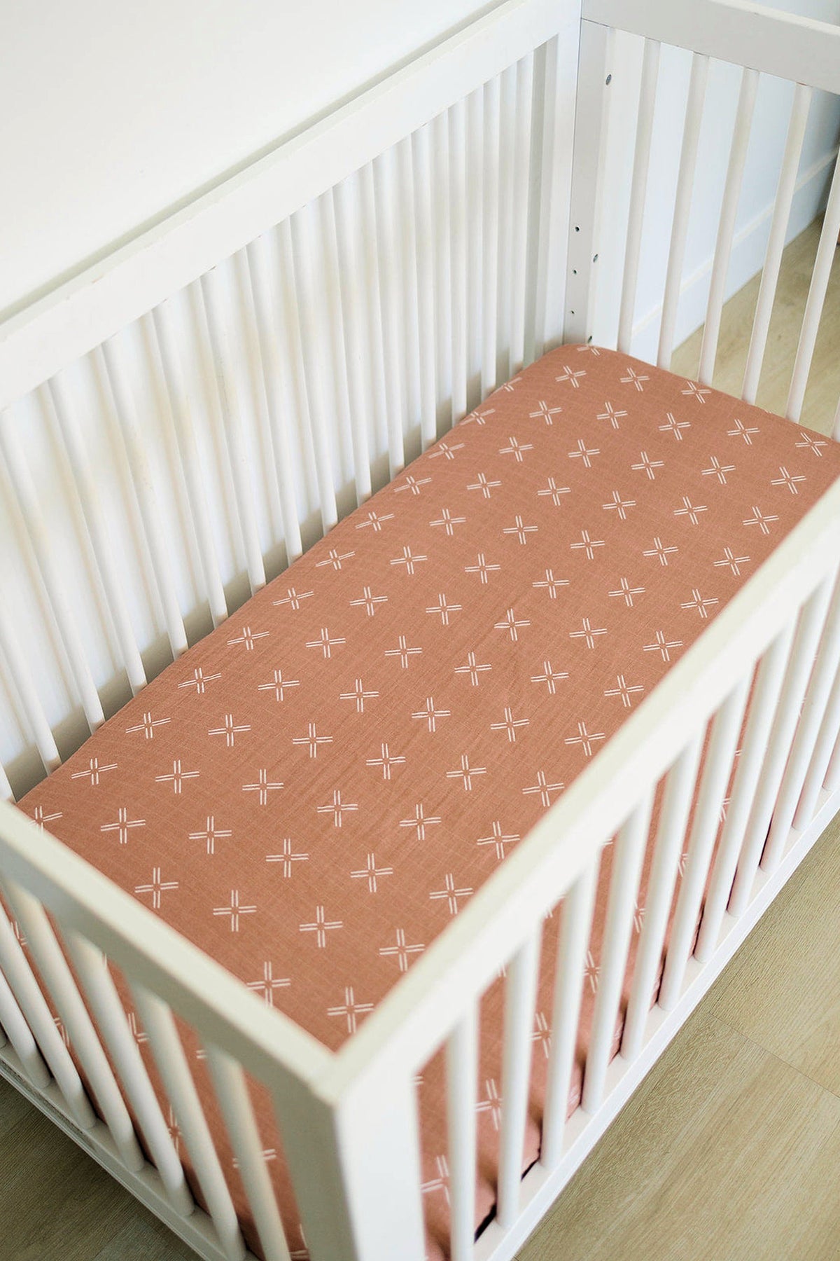 White crib with a peach-colored muslin sheet featuring white cross patterns, set on a light wooden floor.