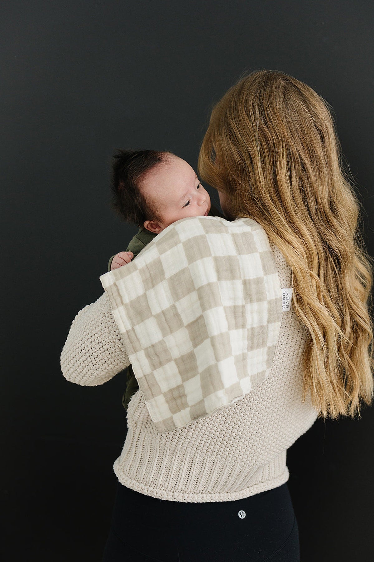 A woman holds a baby with a taupe checkered muslin burp cloth draped over her shoulder.