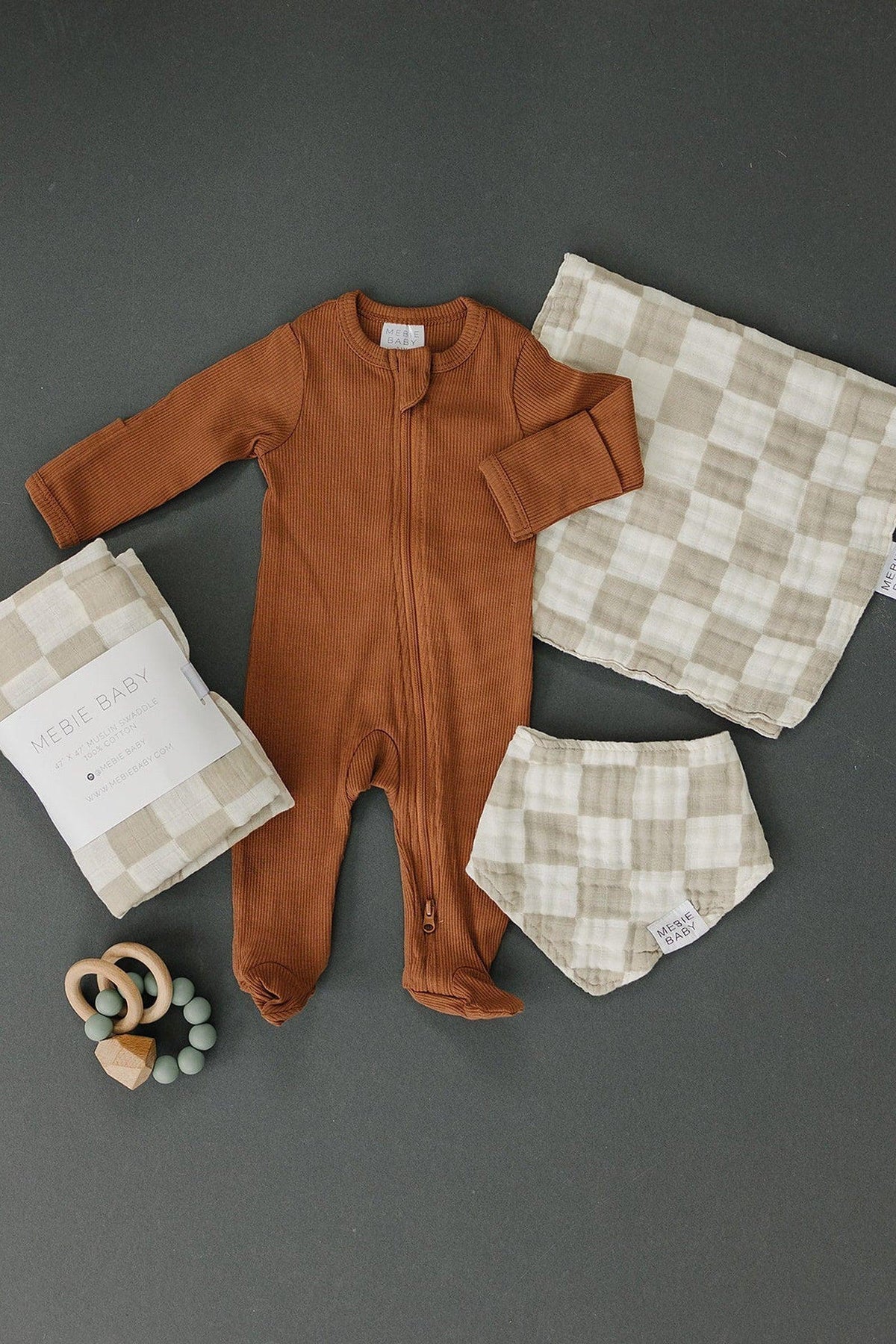 Rust-colored ribbed baby onesie, taupe checkered muslin bib, blanket, and wooden teether on dark background.