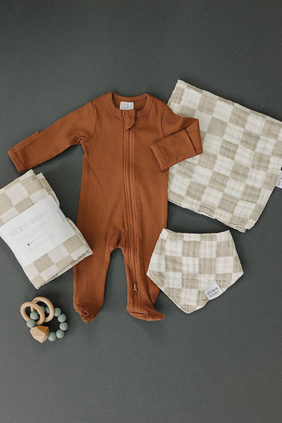 Brown baby romper, taupe checkered burp cloth, matching diaper cover, and wooden teether on a dark background.