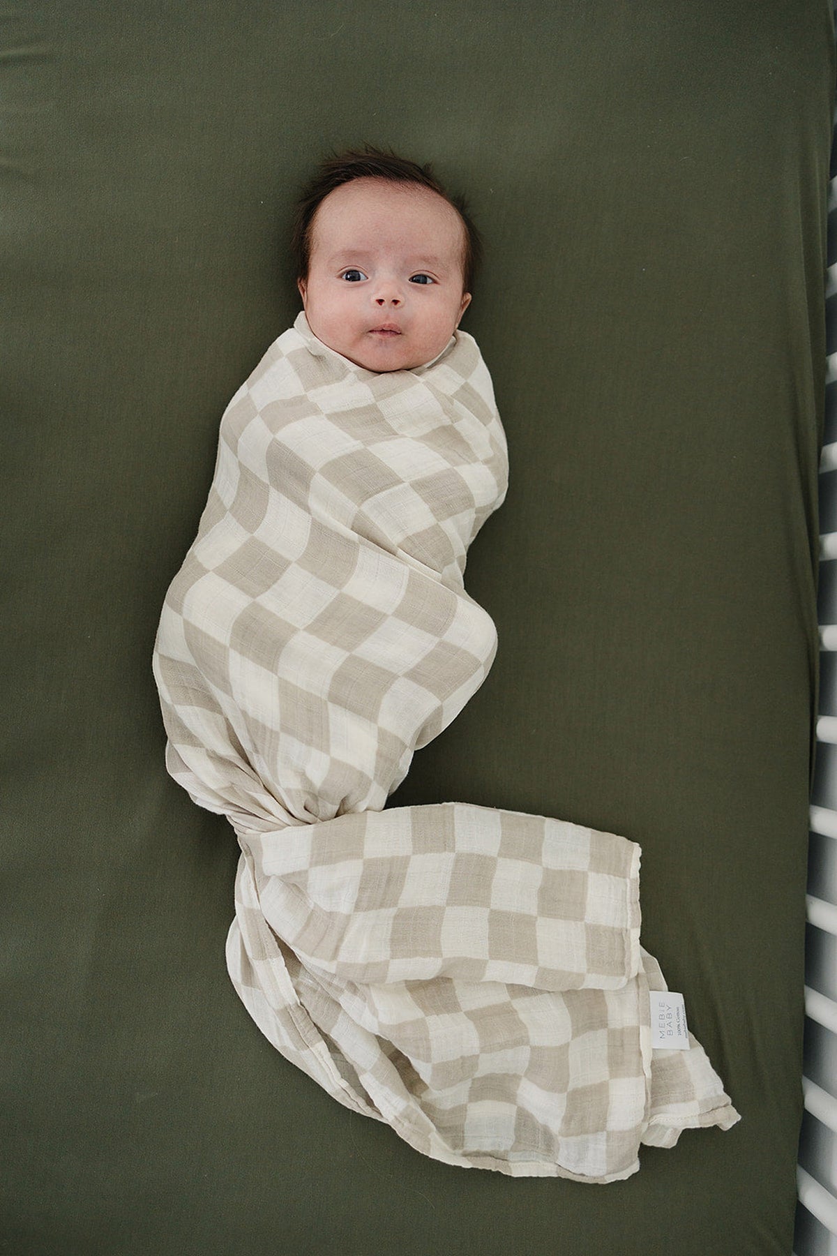A baby is swaddled in a taupe checkered muslin blanket on a dark green surface.