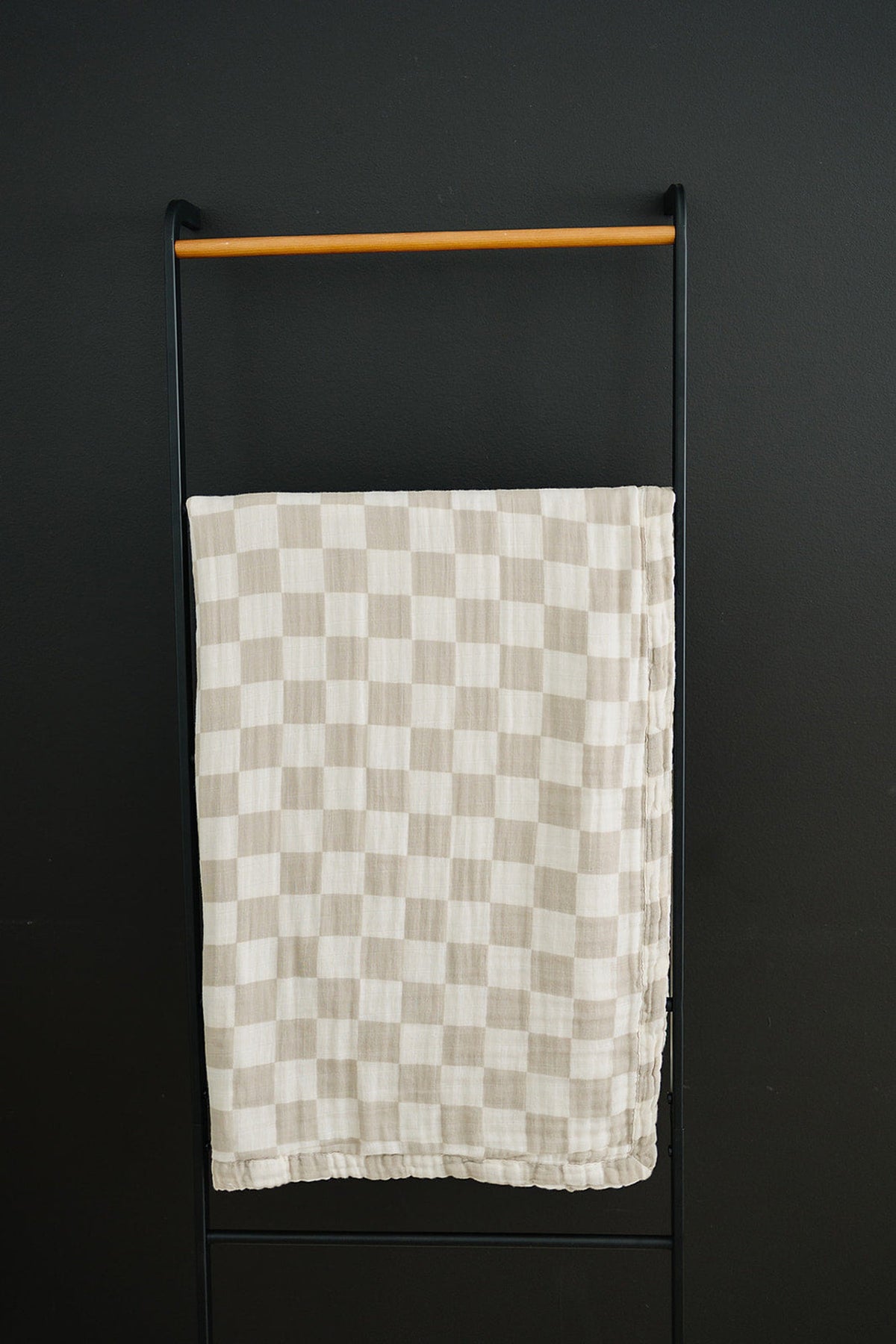 A taupe checkered muslin quilt hangs on a black metal rack against a dark wall.