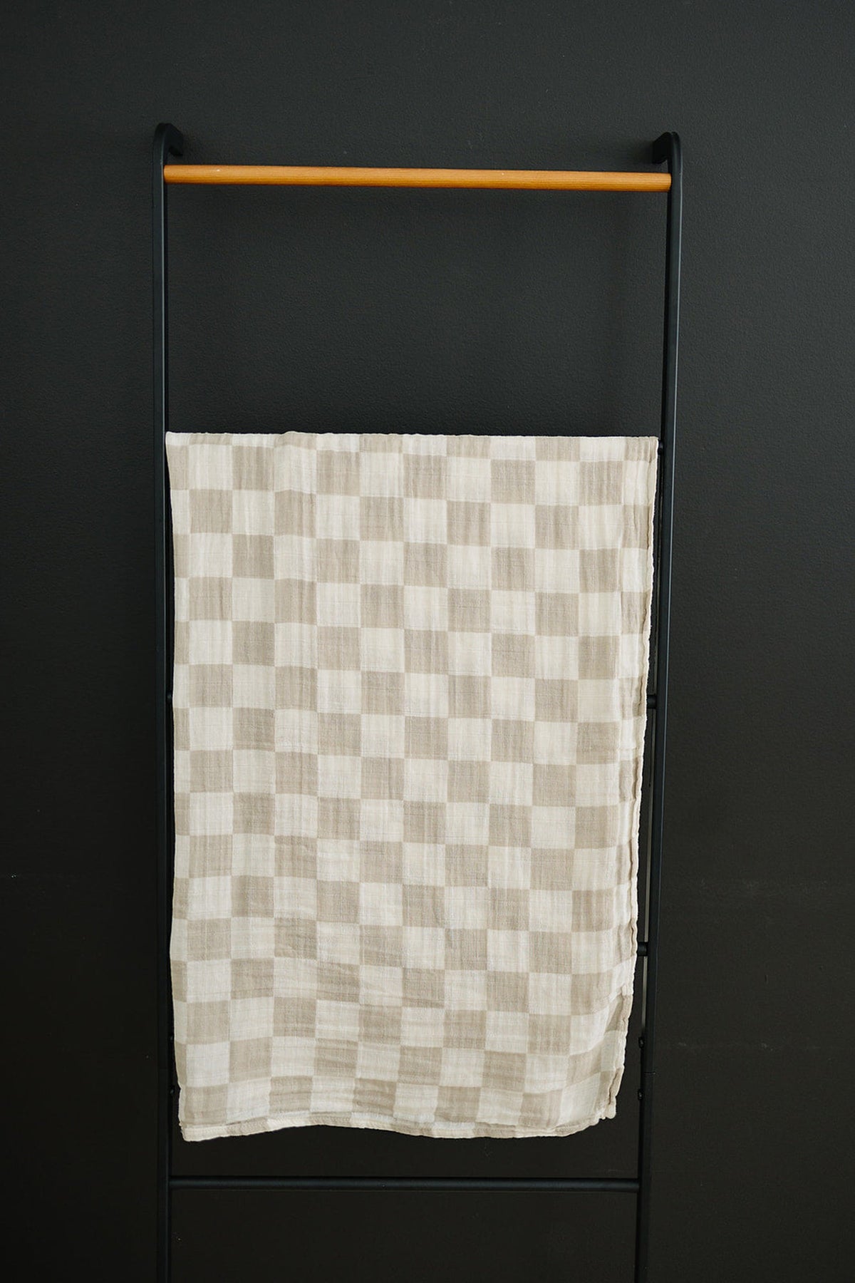 Taupe checkered muslin swaddle blanket hangs on a black metal rack against a dark wall.