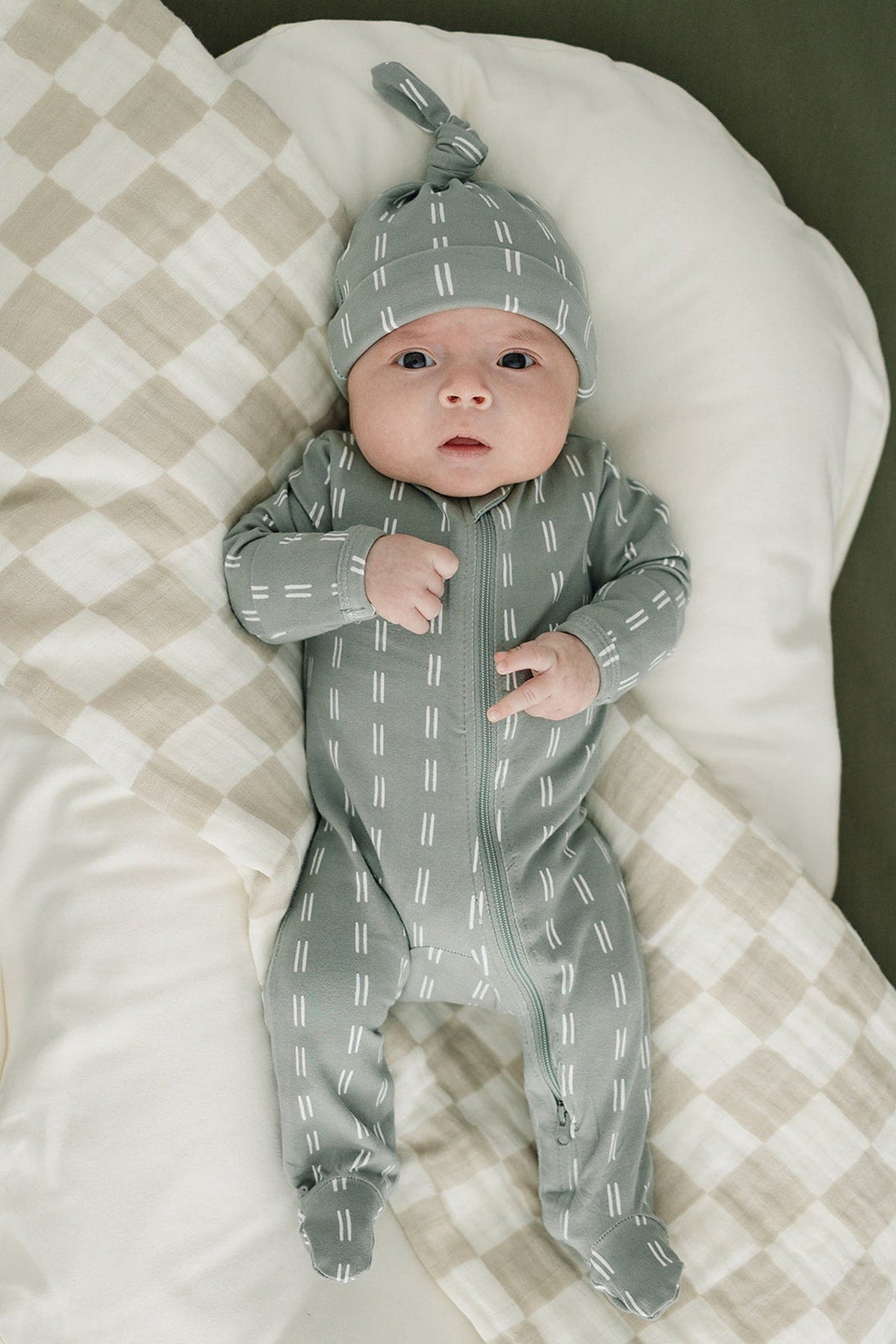 A baby in a gray patterned onesie lies on a taupe checkered muslin quilt, looking up.