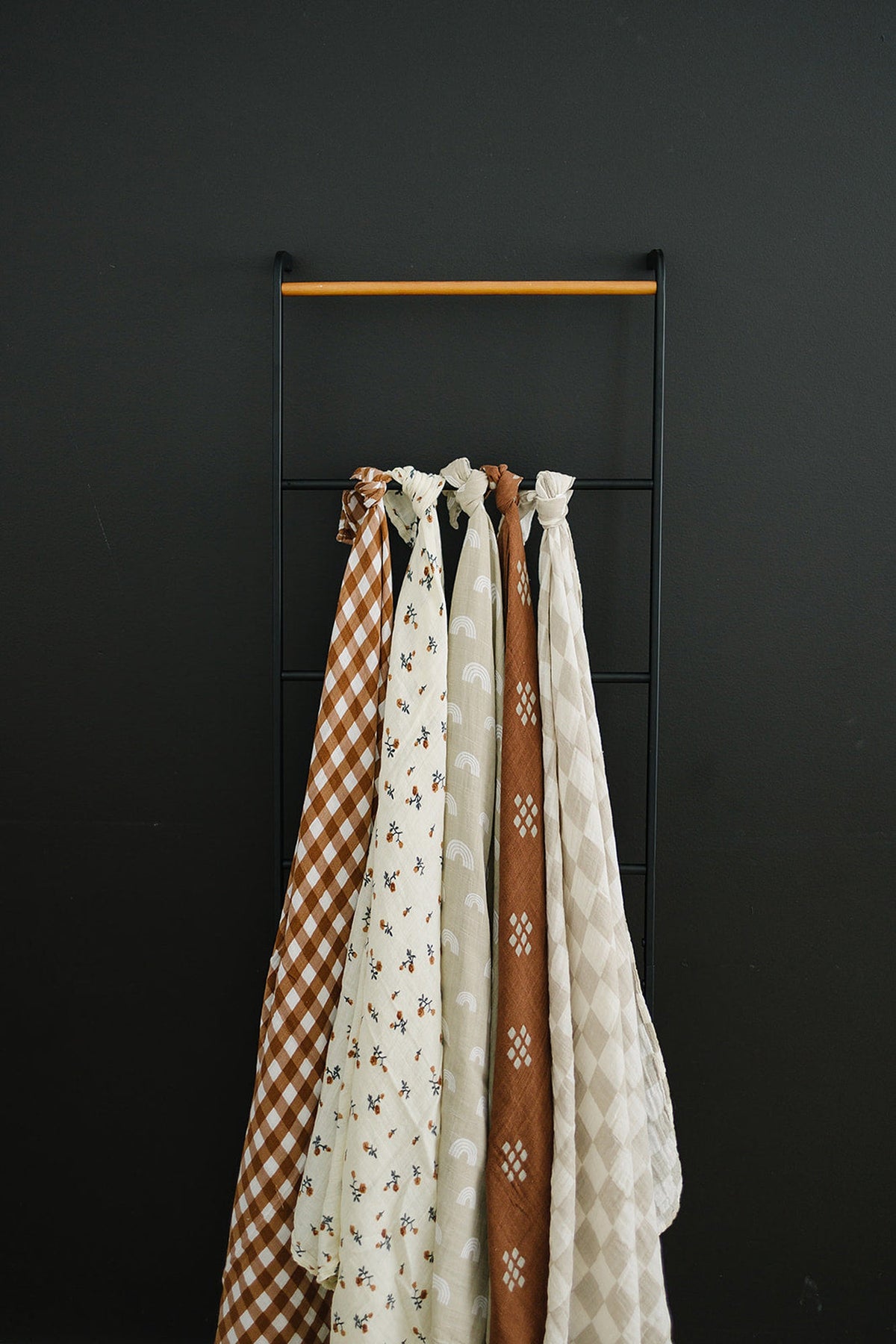 Five patterned muslin swaddle blankets hang on a black ladder against a dark wall, featuring various earthy designs.