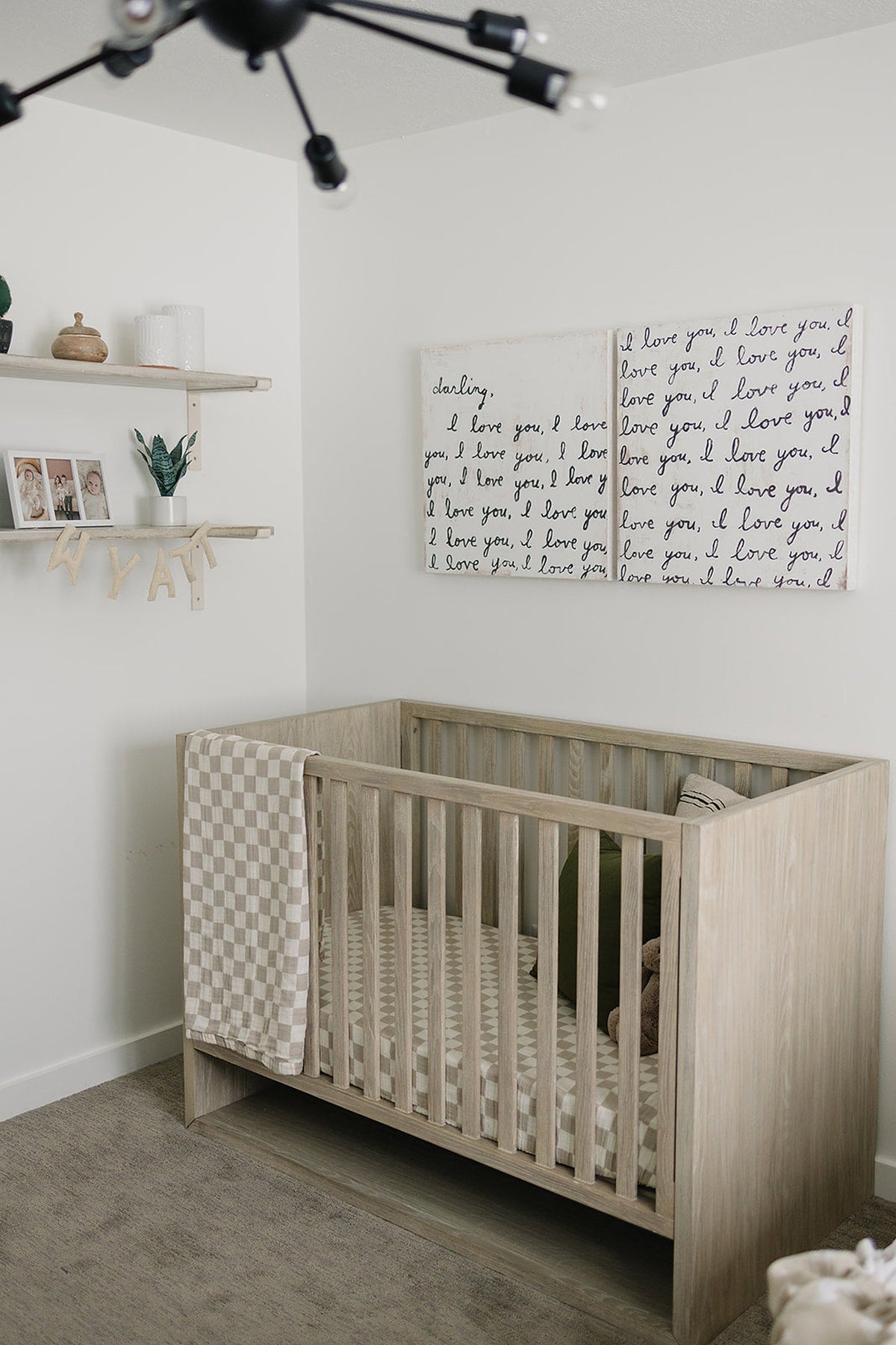 A cozy nursery features a wooden crib with a taupe checkered quilt, decorative wall art, and a shelf.