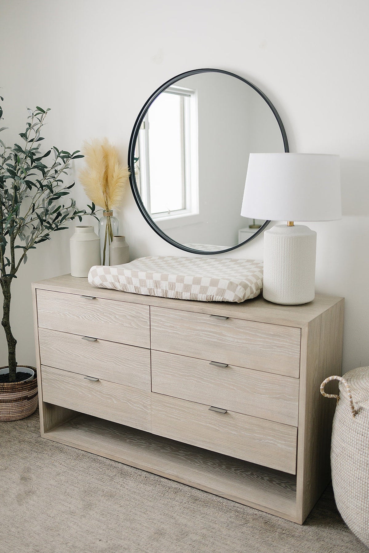 A neutral-toned dresser with six drawers sits beneath a round mirror, featuring a taupe checkered changing pad cover. Decorative plants and a stylish lamp complete the cozy nursery setting.