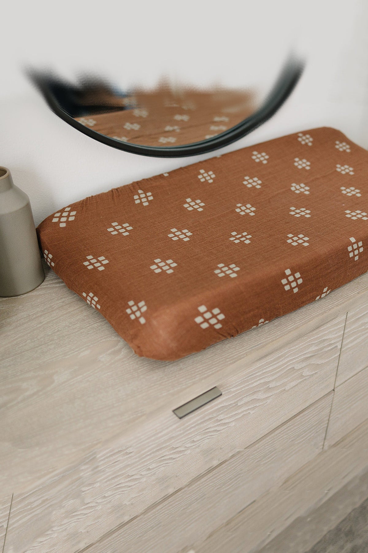 A brown muslin changing pad cover with white patterns is placed on a light wooden dresser.