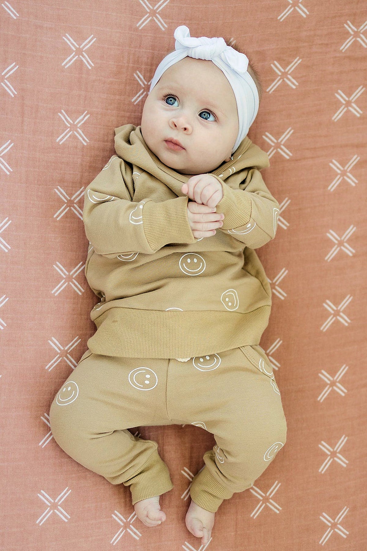 A baby wearing a tan outfit with smiley faces lies on a peach muslin crib sheet with white patterns.
