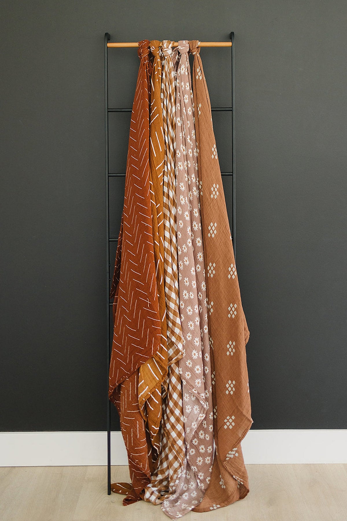 Five patterned muslin blankets in earthy tones hang on a black metal rack against a dark wall.