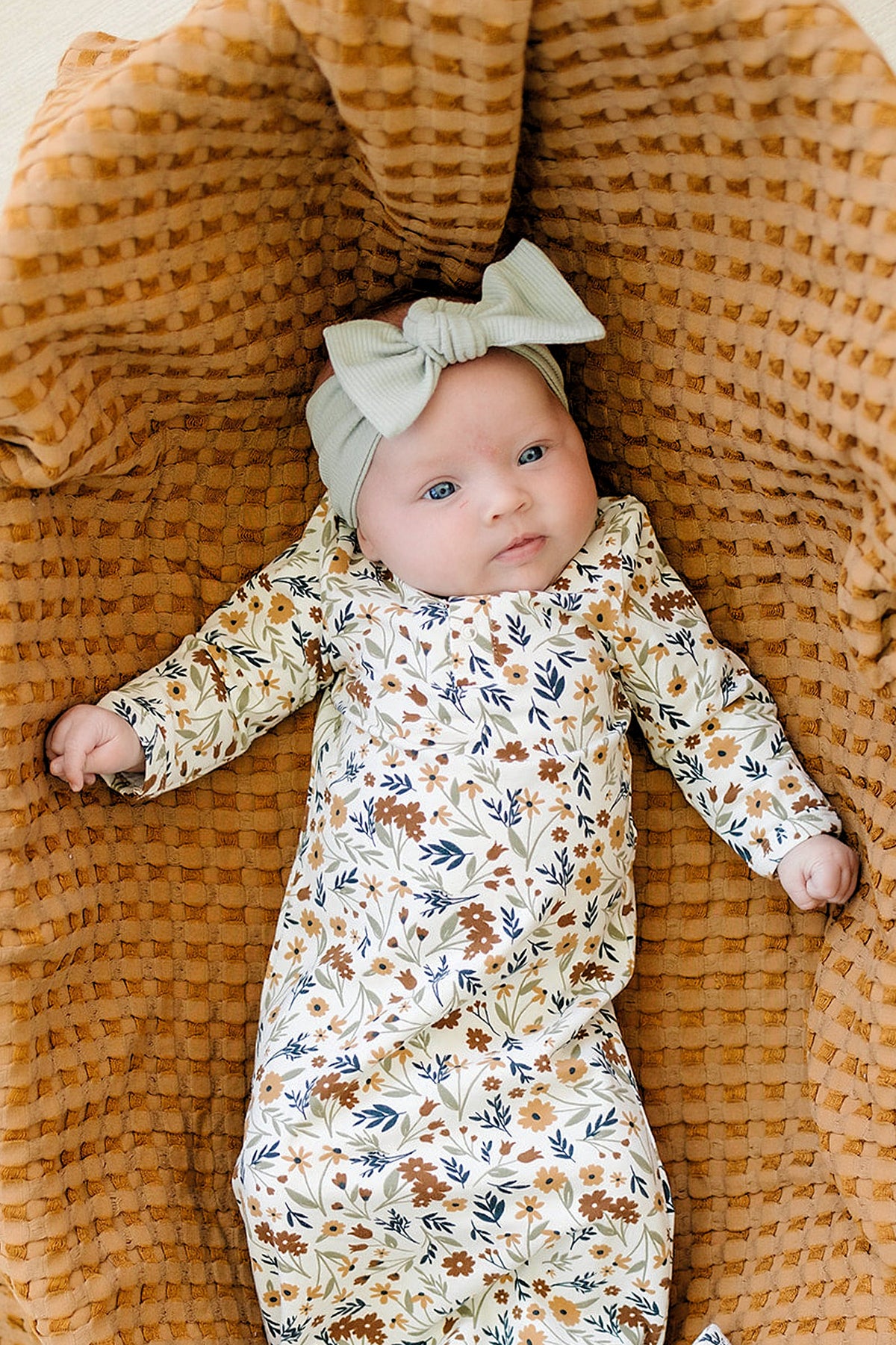 A baby wearing a floral-patterned outfit and sage ribbed head wrap lies on a textured brown blanket.