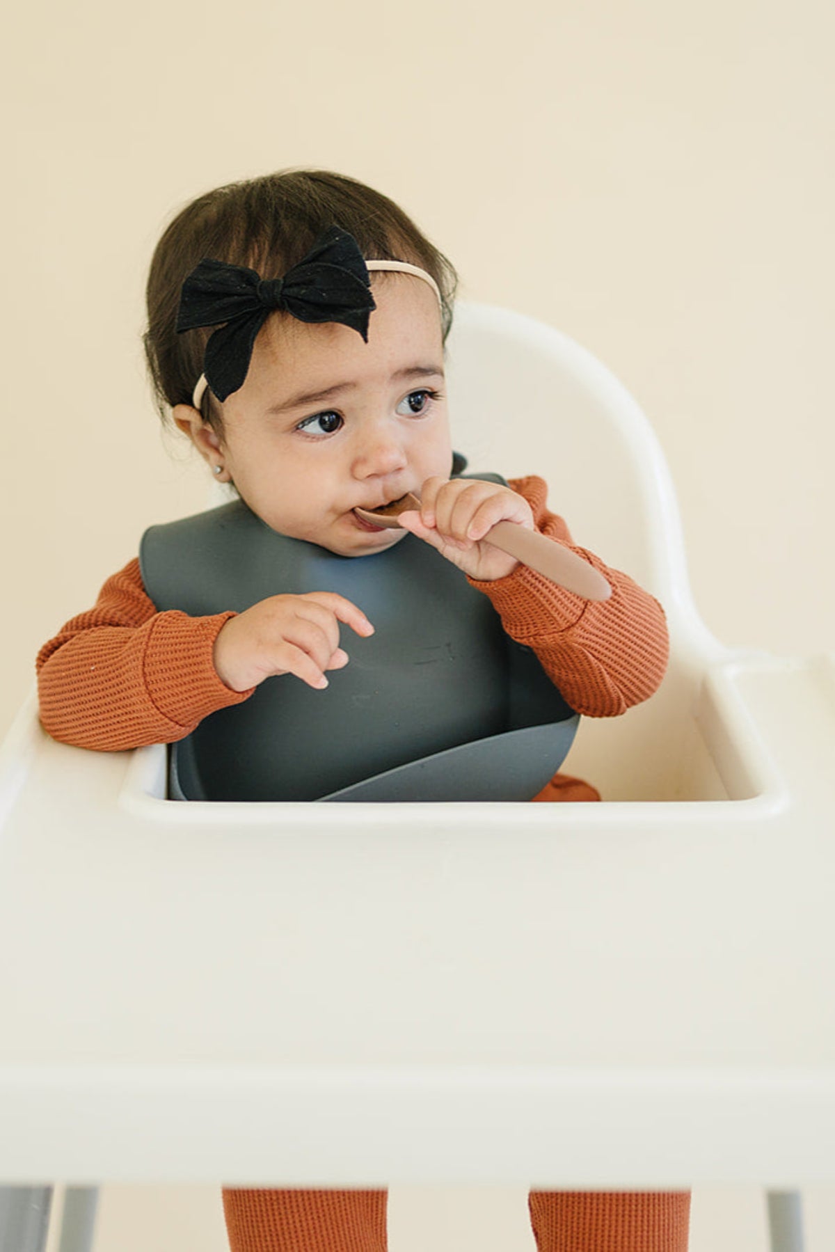 A baby wearing a dark silicone bib sits in a high chair, holding a spoon and wearing a black bow.