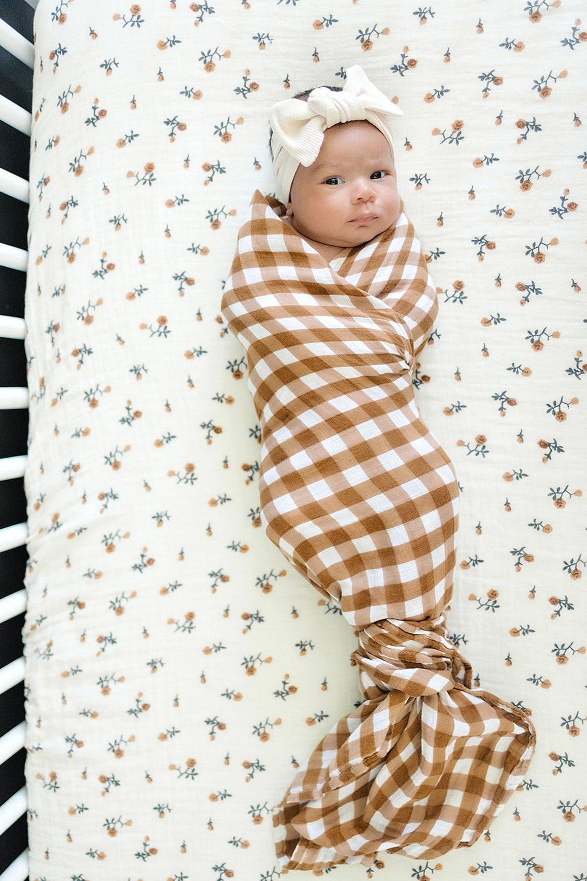 A baby wrapped in a brown and white gingham muslin swaddle, lying on a floral-patterned sheet.
