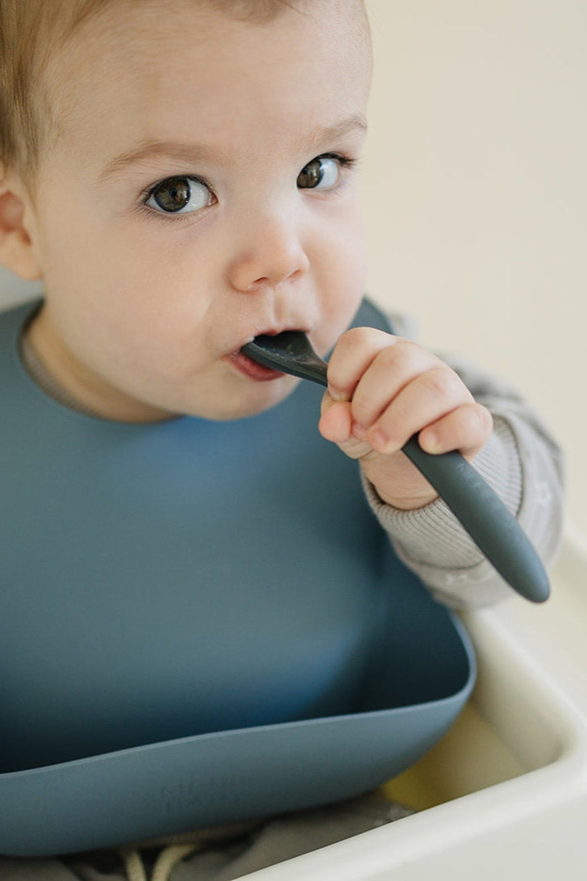 A baby wearing a blue silicone bib is chewing on a dark-colored spoon, sitting in a high chair.