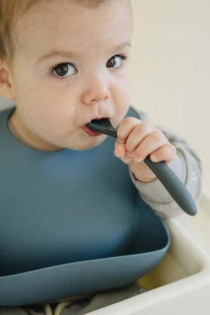 A baby wearing a blue silicone bib is chewing on a dark-colored spoon, sitting in a high chair.