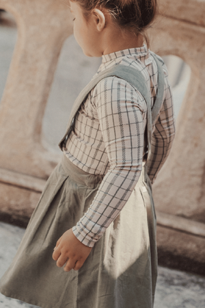 A child stands with their back to the camera, wearing a sage green dress over a checkered long sleeve shirt. The outfit features a playful, stylish design suitable for kids.