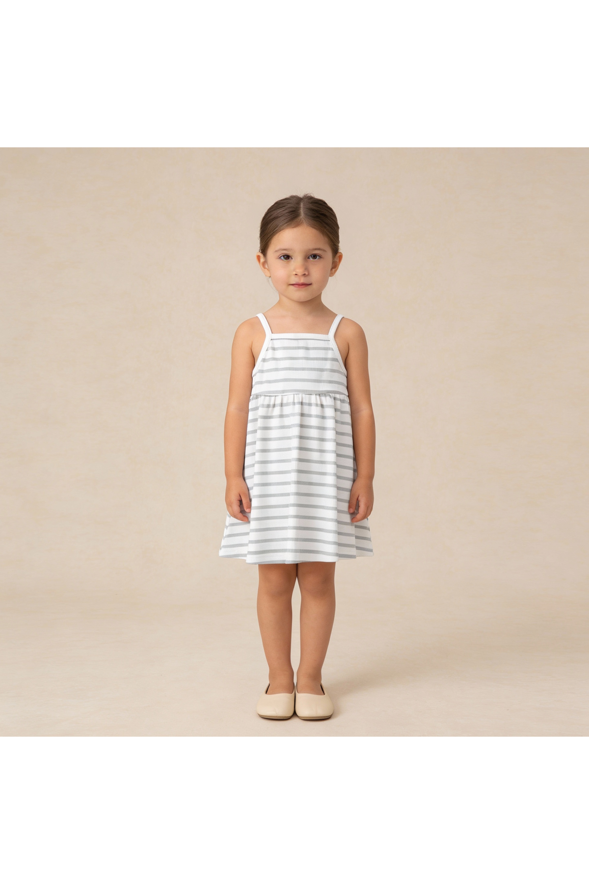 A young girl stands wearing a sleeveless, organic cotton dress featuring light gray and white stripes. The dress has adjustable tie-back straps and a flared skirt, perfect for warm weather.
