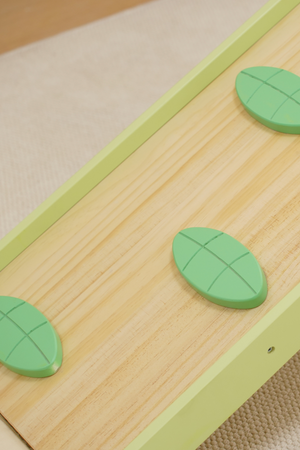 A close-up view of a wooden climbing surface featuring green leaf-shaped handholds. The smooth, sustainably sourced wood enhances safety and playfulness for toddlers in indoor settings.