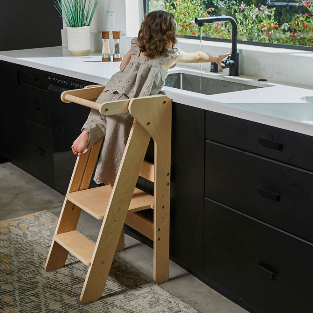 A child climbs the Little Chef Foldable Educational Tower made of light wood, reaching for the kitchen faucet.