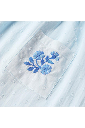 Close-up view of a light blue organic cotton fabric featuring a delicate embroidered pocket with floral design. The texture is airy with a subtle dobby weave, perfect for sunny days.