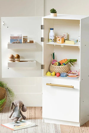 A white wooden play refrigerator is open, showcasing colorful toy food items like fruits, vegetables, and dairy products. A cozy stuffed animal sits on the floor beside the fridge.