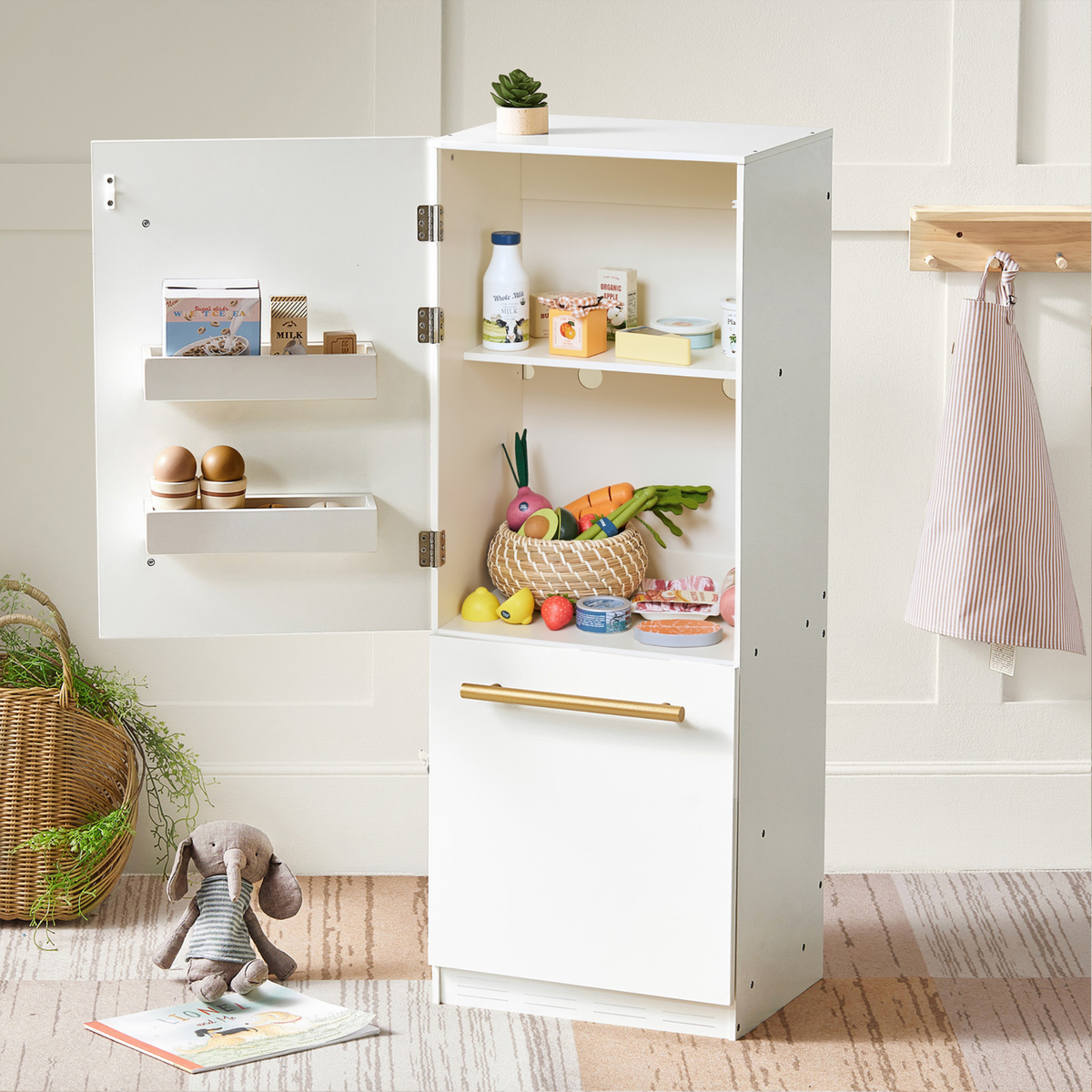 A white wooden play refrigerator stands open, showcasing colorful toy fruits and vegetables on its shelves. A plush bunny sits nearby on a soft, textured rug, enhancing the playful setting.