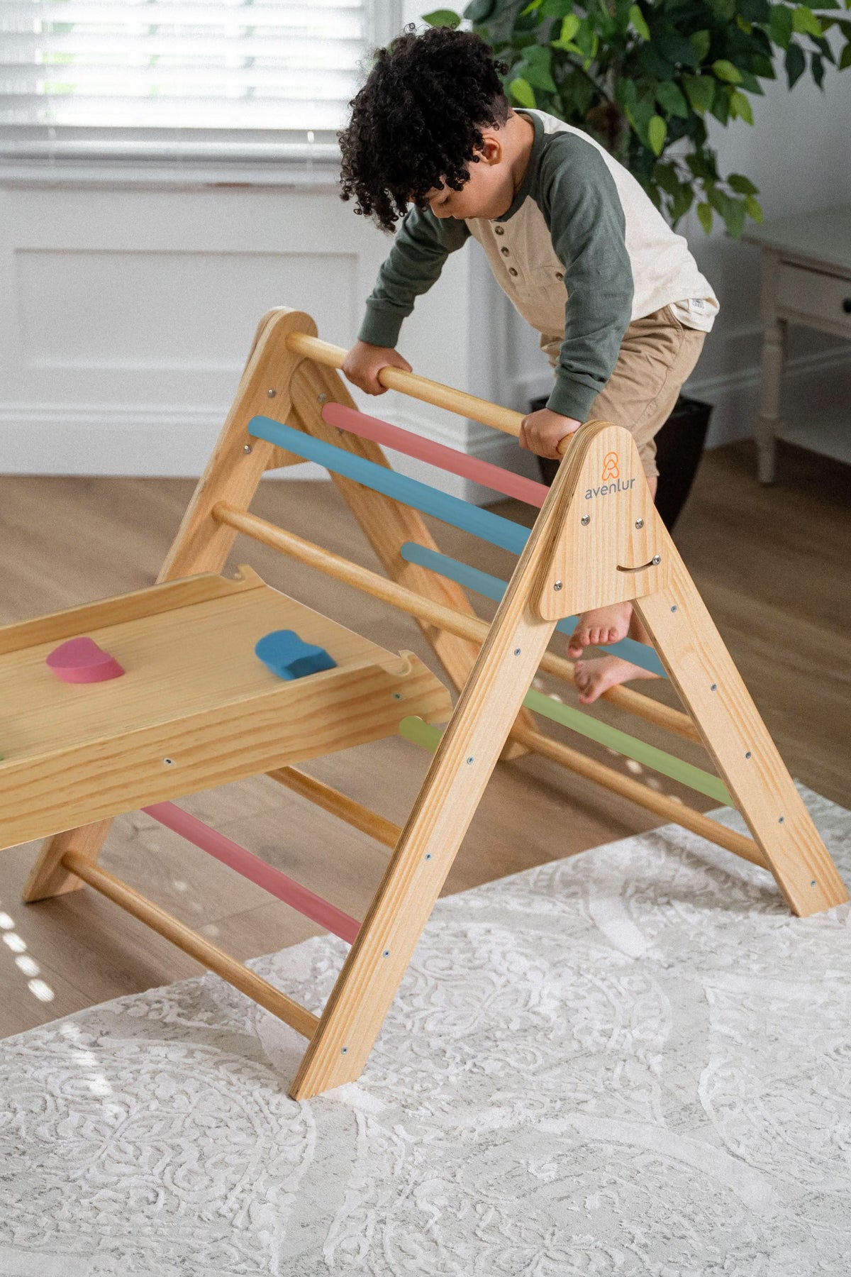 A child climbs on a natural wood Pikler triangle playset with colorful grips. The indoor setting features light flooring and bright natural light streaming through a window.