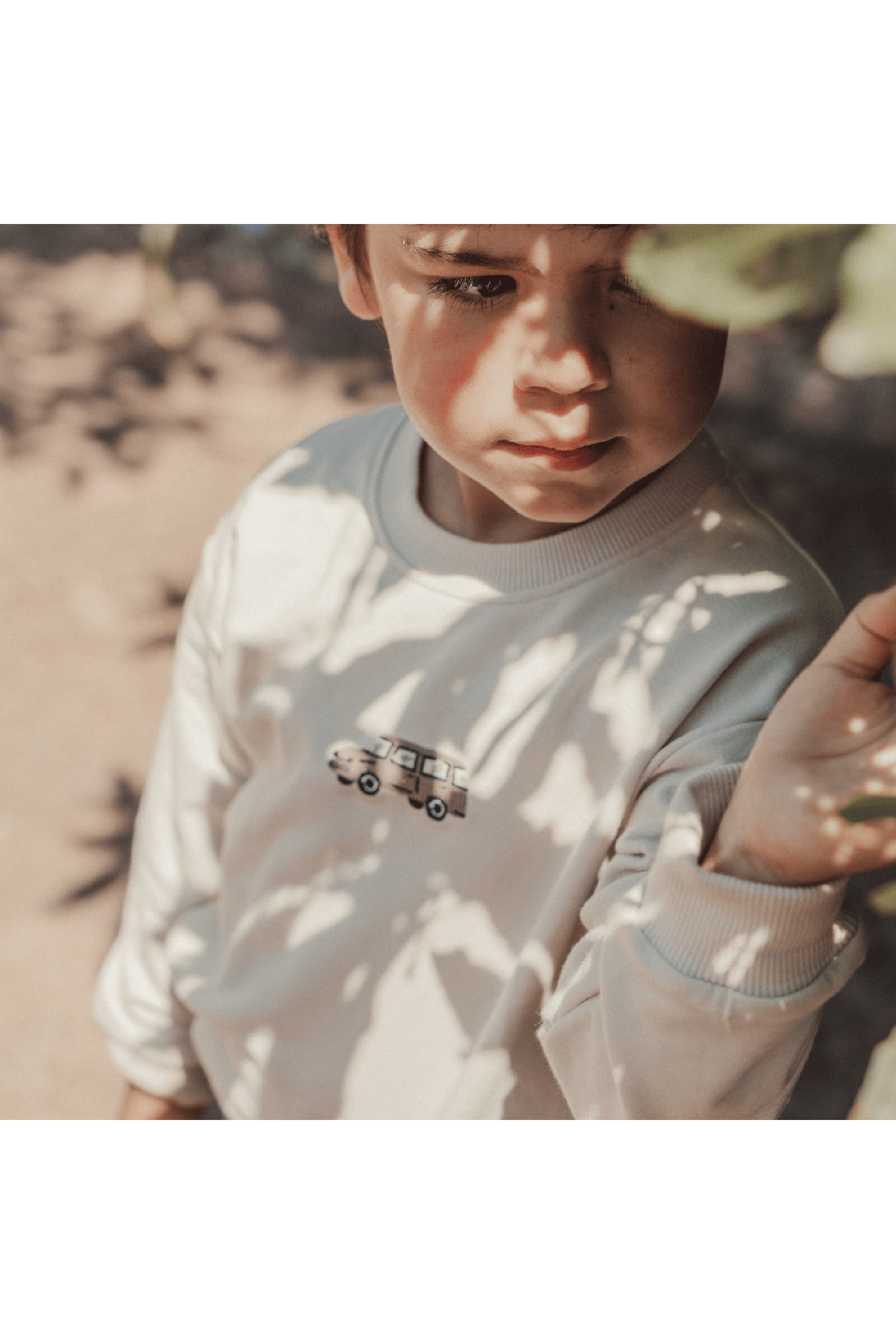 A young child wearing a light-colored sweatshirt with a vintage car embroidery stands amidst leafy shadows. The soft cotton fabric is perfect for cozy outdoor adventures.