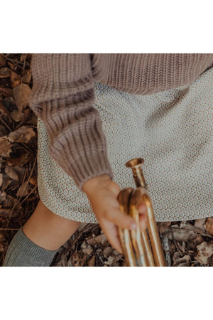 A child sits on a bed of leaves, wearing a light-colored textured skirt and a cozy sweater. They hold a brass instrument, highlighting a playful autumn setting.