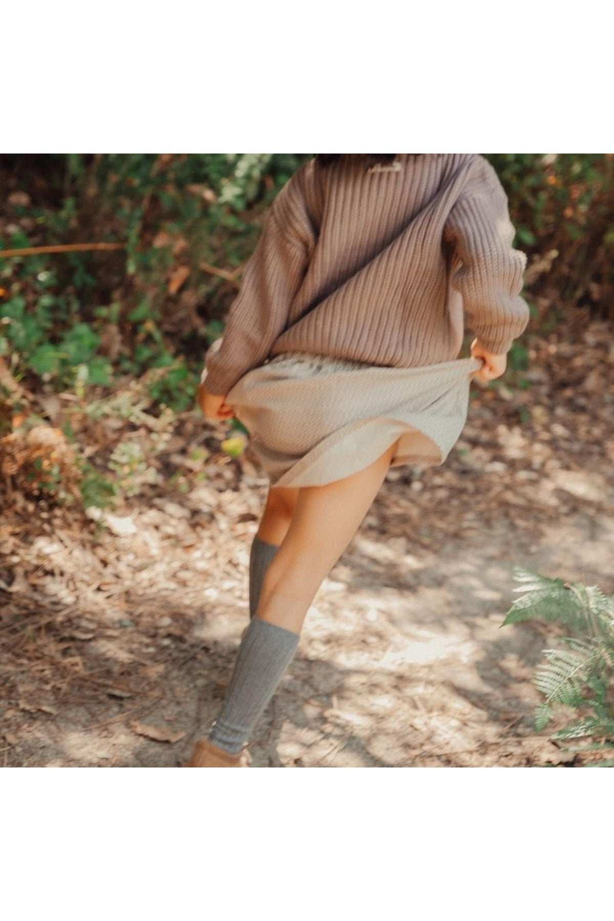 A girl in a cozy, oversized beige sweater twirls, revealing a light-colored skirt. She wears knee-high gray socks, surrounded by lush greenery and a natural, earthy trail.