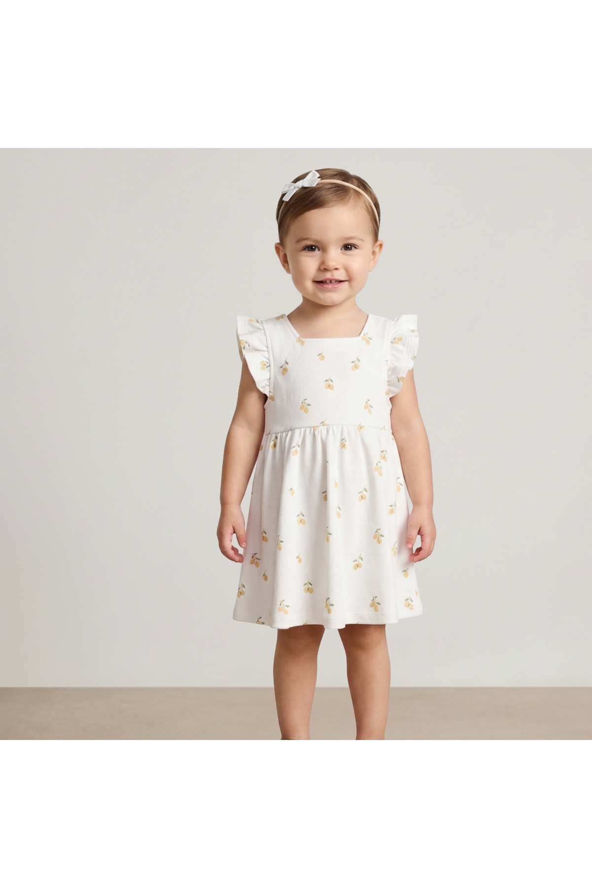 A young girl smiles while wearing a soft, white dress adorned with a cheerful lemon print. The dress features ruffled sleeves, perfect for warm weather and playful activities.