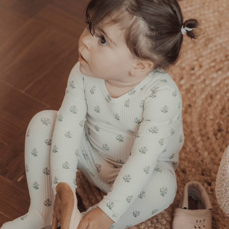 A toddler sits on a textured rug, wearing a light-colored, ribbed outfit adorned with small floral patterns. The child looks curiously to the side, showcasing a playful and cozy atmosphere.