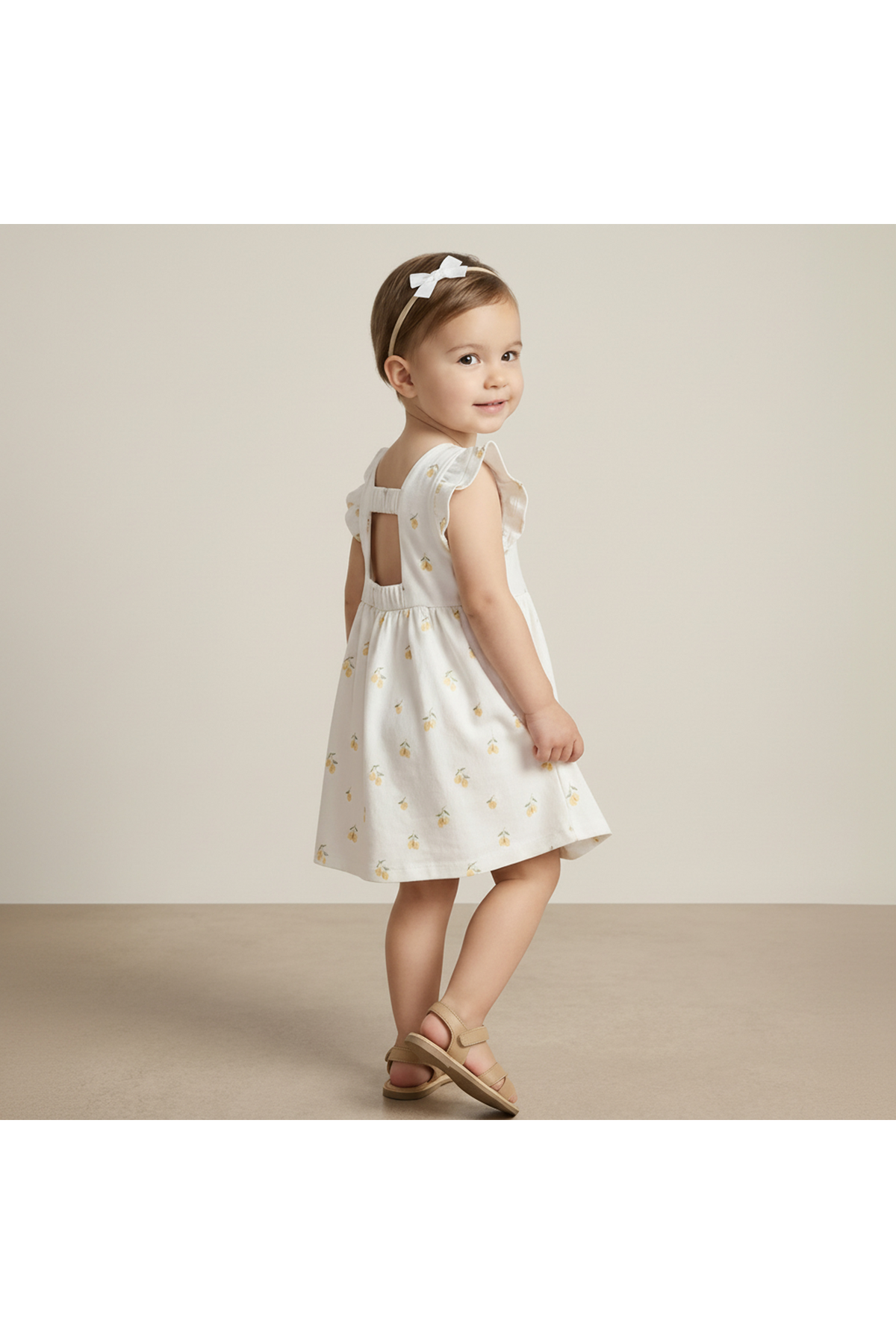 A young child models a white dress with a lemon print and a cutout back. The dress is made from soft, breathable organic cotton, perfect for warm weather play.